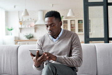 Serious african american man holding digital tablet computer sitting on couch at home. Young adult black guy using apps on tab technology device reading, ordering online or browsing internet.