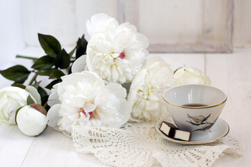 white cup with flower pattern with tea , sweets on the saucer and white peonies lying next to the white wooden background