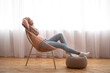 © Prostock-studio - Side view of happy Muslim woman in hijab relaxing in armchair, resting her feet on knitted pouf, copy space