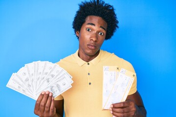 Canvas Print - Handsome african american man with afro hair holding boarding pass and bunch of dollars clueless and confused expression. doubt concept.