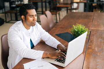 Poster - Focused young African male doctor in white coat working typing on modern laptop computer looking on display screen of monitor, sitting at desk at hospital. Concept of medicine and health care.