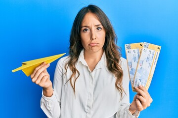 Wall Mural - Young brunette woman holding paper plane and boarding pass puffing cheeks with funny face. mouth inflated with air, catching air.