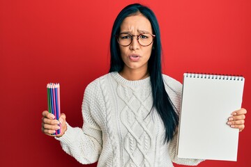 Sticker - Beautiful hispanic woman holding canvas book and colored pencils in shock face, looking skeptical and sarcastic, surprised with open mouth