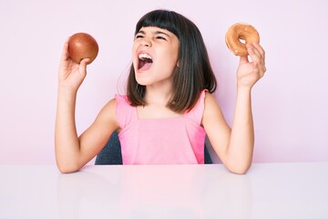 Wall Mural - Young little girl with bang holding red apple and donut sitting on the table angry and mad screaming frustrated and furious, shouting with anger. rage and aggressive concept.