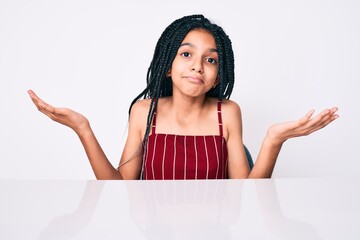 Canvas Print - Young african american girl child with braids wearing casual clothes sitting on the table clueless and confused expression with arms and hands raised. doubt concept.