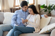 © fizkes - Online together. Happy loving young family couple sitting on couch browsing internet or social networks using cellphone, making video call, chatting with friends, watching cute photos on gadget screen