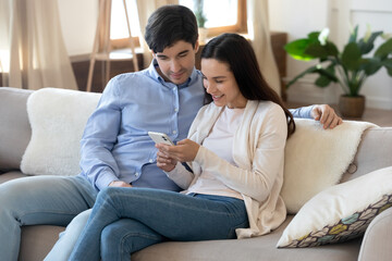 Sticker - Online together. Happy loving young family couple sitting on couch browsing internet or social networks using cellphone, making video call, chatting with friends, watching cute photos on gadget screen