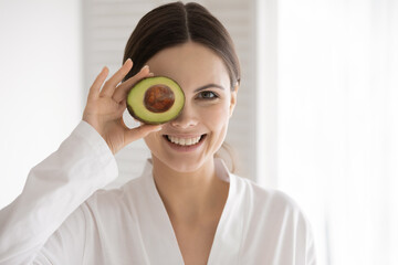 Wall Mural - Benefits of proper nutrition. Joyful young female with healthy skin and white beautiful smile posing for portrait looking at camera hiding eye behind avocado slice, recommends to use natural cosmetics