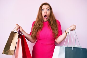 Canvas Print - Young beautiful redhead woman holding shopping bags afraid and shocked with surprise and amazed expression, fear and excited face.