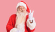 Old senior man with grey hair and long beard wearing traditional santa claus costume smiling with happy face winking at the camera doing victory sign with fingers. number two.