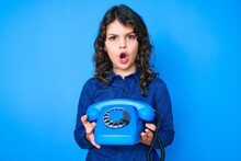 Cute Hispanic Boy With Long Hair Holding Vintage Telephone In Shock Face, Looking Skeptical And Sarcastic, Surprised With Open Mouth