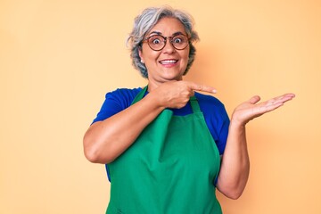 Sticker - Senior hispanic woman wearing apron and glasses amazed and smiling to the camera while presenting with hand and pointing with finger.