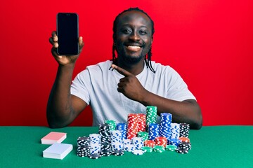 Poster - Handsome young black man playing poker holding smartphone smiling happy pointing with hand and finger