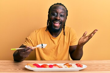 Sticker - Handsome young black man eating sushi sitting on the table celebrating achievement with happy smile and winner expression with raised hand