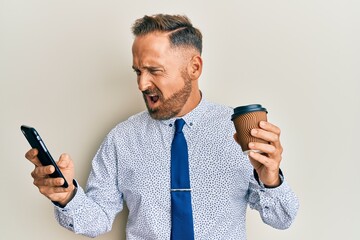 Wall Mural - Handsome middle age business man using smartphone and drinking a cup of coffee angry and mad screaming frustrated and furious, shouting with anger. rage and aggressive concept.