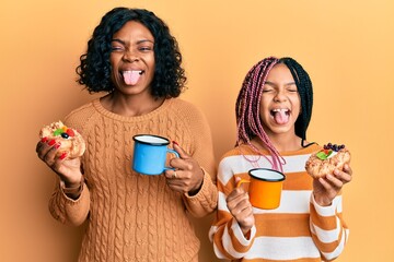 Sticker - Beautiful african american mother and daughter holding sweet pastries and drinking coffee sticking tongue out happy with funny expression.