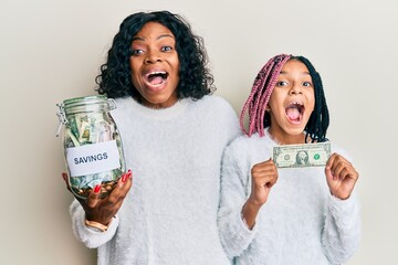 Wall Mural - Beautiful african american mother and daughter holding jar with savings and 1 dollar banknote celebrating crazy and amazed for success with open eyes screaming excited.