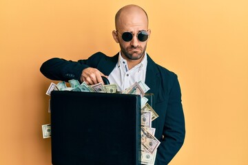 Hispanic bald business man wearing sunglasses holding briefcase full of dollars sticking tongue out happy with funny expression.