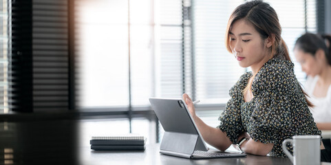 Young attractive asian woman working on tablet computer while sitting in modern office room with her friend.