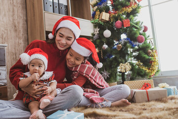 Wall Mural - parent and two little children having fun and playing together near christmas tree indoors. merry christmas and happy holidays. cheerful mom and her cute daughters girls exchanging gifts.