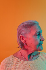 Wall Mural - Profile of serious concentrated old lady in light knitted clothes isolated over bright orange background. Pearl necklace on her neck. Close up portrait.