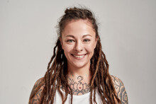 Close Up Portrait Of Young Tattooed Caucasian Woman With Dreadlocks Wearing White Shirt, Smiling At Camera While Posing Isolated Over Grey Background