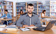© JackF - Focused efficient friendly guy preparing for exam in college library, reading book and using laptop for searching information