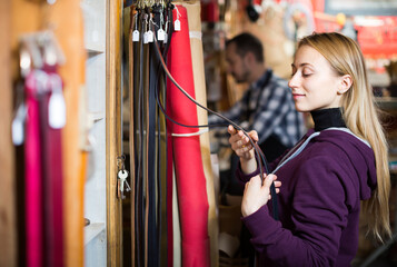 Young russian woman choosing new belt in leather workshop.