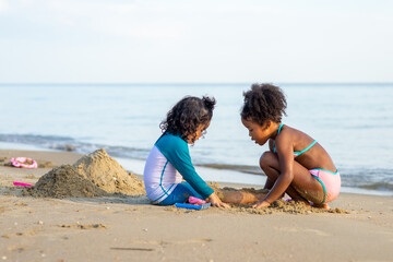 Adorable little mixed race girl with Asian girl friend in swimsuit sitting on the beach and playing sand with beach toys together. Two cute child girl relax and having fun on summer holiday vacation.