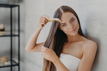 Young Brunette Woman Brushes Hair With Comb After Taking Shower And Applying Hair Care Mask, Wears Minimal Makeup, Has Healthy Glowing Skin After Hygienic Procedures, Stands Wrapped In Towel