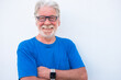 Portrait close-up of a smiling senior man with white beard on white background wearing eyeglasses and tshirt in blue color. Positive retiree person with crossed arms looking at camera