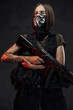 Armed with ak74 rifle and weared with mask stylish female mercenary poses in dark background.