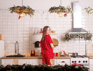 Woman in pyjamas smiling wide and calling to taste festive food. Cooking dinner at christmas eve in decorated kitchen. Home interior with xmas decoration. Preparing for new year celebration