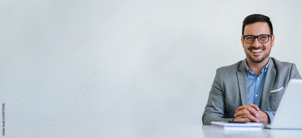 Banner panorama portrait of young smiling cheerful businessman manager entrepreneur with eyeglasses sitting at desk in office or at home looking directly at camera hands fingers crossed copy space - obrazy, fototapety, plakaty 