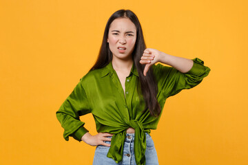 Wall Mural - Displeased dissatisfied disgusted young brunette asian woman wearing casual basic green shirt standing showing thumb down looking camera isolated on bright yellow colour background studio portrait.