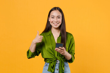 Wall Mural - Smiling pretty young brunette asian woman wearing basic green shirt standing using mobile cell phone typing sms message showing thumb up isolated on bright yellow colour background, studio portrait.