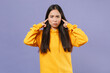 © ViDi Studio - Dissatisfied displeased young brunette asian woman wearing casual basic yellow hoodie standing covering ears with fingers looking camera isolated on pastel violet colour background studio portrait.