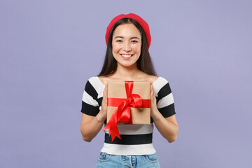 Wall Mural - Smiling cute young brunette asian woman 20s in striped t-shirt red beret standing hold red present box with gift ribbon bow looking camera isolated on pastel violet colour background studio portrait.