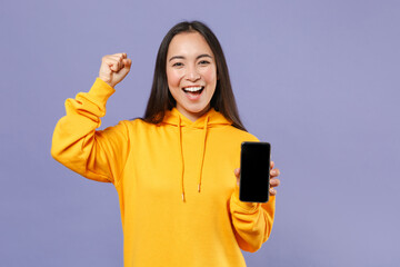 Wall Mural - Joyful young brunette asian woman 20s in basic yellow hoodie standing hold mobile cell phone with blank empty screen doing winner gesture isolated on pastel violet colour background, studio portrait.