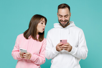 Wall Mural - Smiling curious young couple two friends man woman 20s wearing white pink casual hoodie using mobile cell phone typing sms message isolated on blue turquoise colour wall background studio portrait.