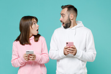 Wall Mural - Shocked young couple two friends man woman 20s wearing white pink casual hoodie using mobile cell phone typing sms message looking at each other isolated on blue turquoise background studio portrait.