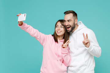 Wall Mural - Cheerful young couple friends man woman 20s in white pink casual hoodie doing selfie shot on mobile phone showing victory sign hugging isolated on blue turquoise colour background studio portrait.