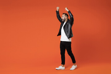 Wall Mural - Full length side view of shocked young bearded man in basic white t-shirt black leather jacket standing expressive gesticulating with rising hands isolated on orange colour background studio portrait.