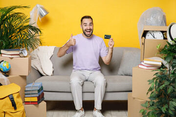 Wall Mural - Excited young owner man hold credit bank card showing thumb up sits in living room on sofa at home household unpacking stuff rents flat isolated on yellow wall. Relocation moving in apartment concept.