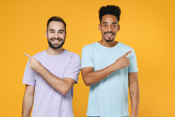 Wall Mural - Cheerful funny young friends european african american men 20s wearing casual violet blue t-shirts pointing index fingers aside looking camera isolated on yellow colour background studio portrait.