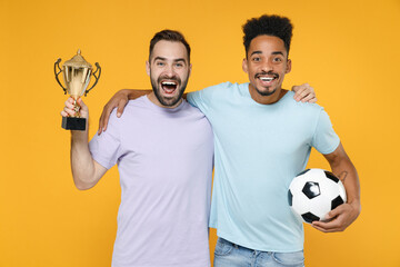 Wall Mural - Excited young friends european african american men football fans in casual t-shirts cheer up support favorite team with soccer ball golden cup hugging isolated on yellow background studio portrait.