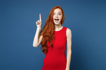 Wall Mural - Excited amazed young redhead woman 20s wearing bright red elegant evening dress standing holding index finger up with great new idea looking camera isolated on blue color background studio portrait.