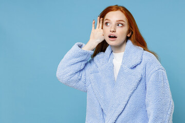 Wall Mural - Shocked curious young redhead woman 20s in warm fur coat standing try to hear you overhear listening intently with hand near ear looking aside isolated on pastel blue color background studio portrait.