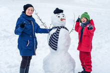 A Grandmother With A Grandson Granddaughter Is Playing With A Snowman. Family Winter Games With Snowballs. Outdoor Activities In The Winter. Family Leisure. Elderly Woman And Child Together