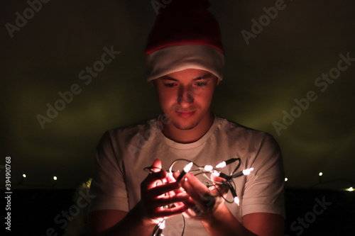 Fotomural Young man enjoys the atmosphere of a real European Christmas and smiles over small lights that he fixes on a Christmas tree